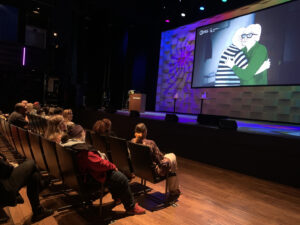 Yleisöä istumassa penkeissä kahdessa rivissä ja katsomassa isoa screeniä, jossa animaatiossa kaksi ihmistä halaa toisiaan.