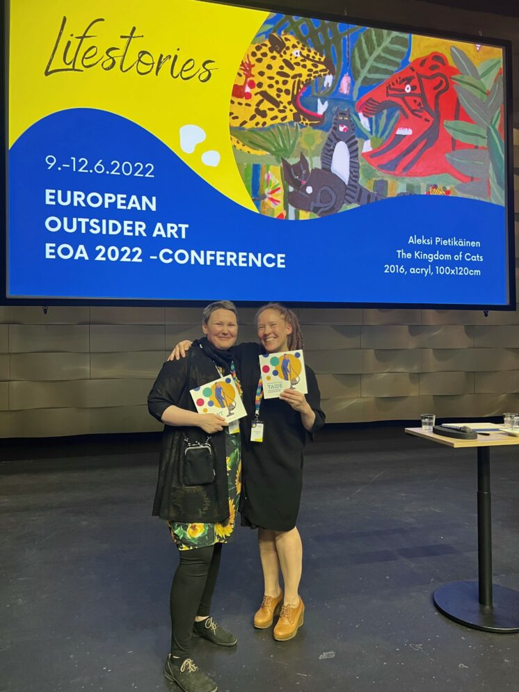 Kaksi henkilöä seisoo lavalla kirjat kädessään ison screenin edessä, jossa on Europen Outsider Art -kongressigrafiikka.