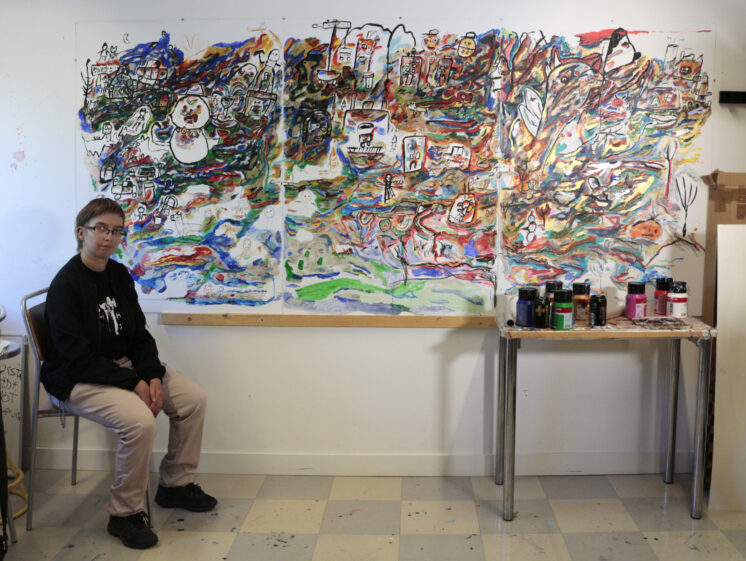 Riina Noro istuu ison maalauksensa Ison Pöön vierellä. Pöydällä on paljon maalaustarvikkeita.
